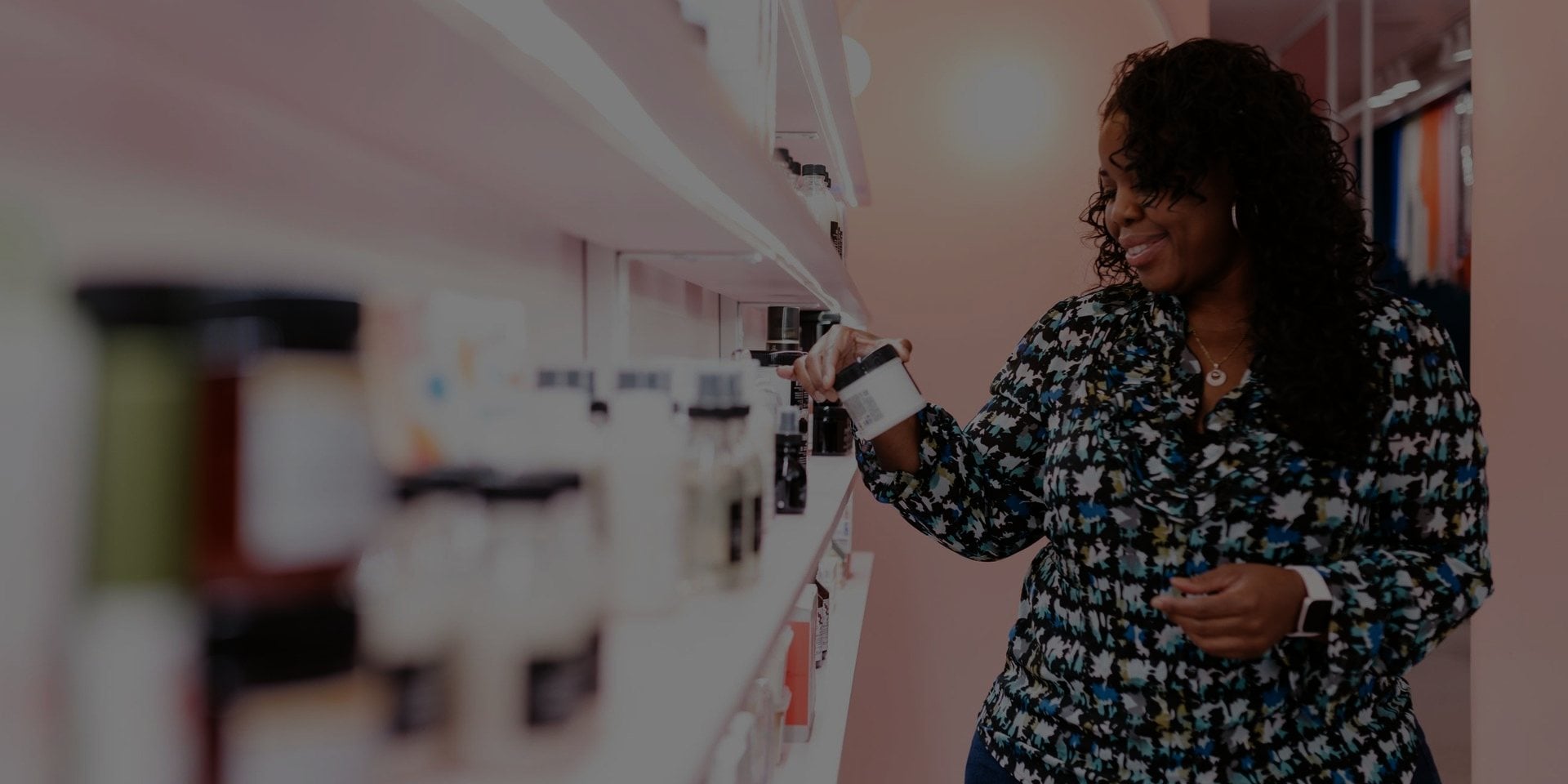 A shopper at a lit wall of beauty product, reading a label.