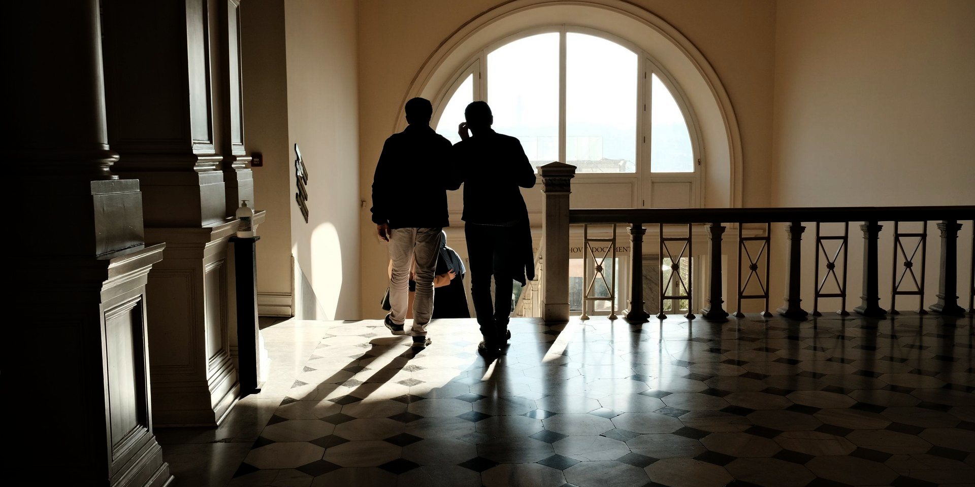 Two people crossing a grand hall under an arched window.